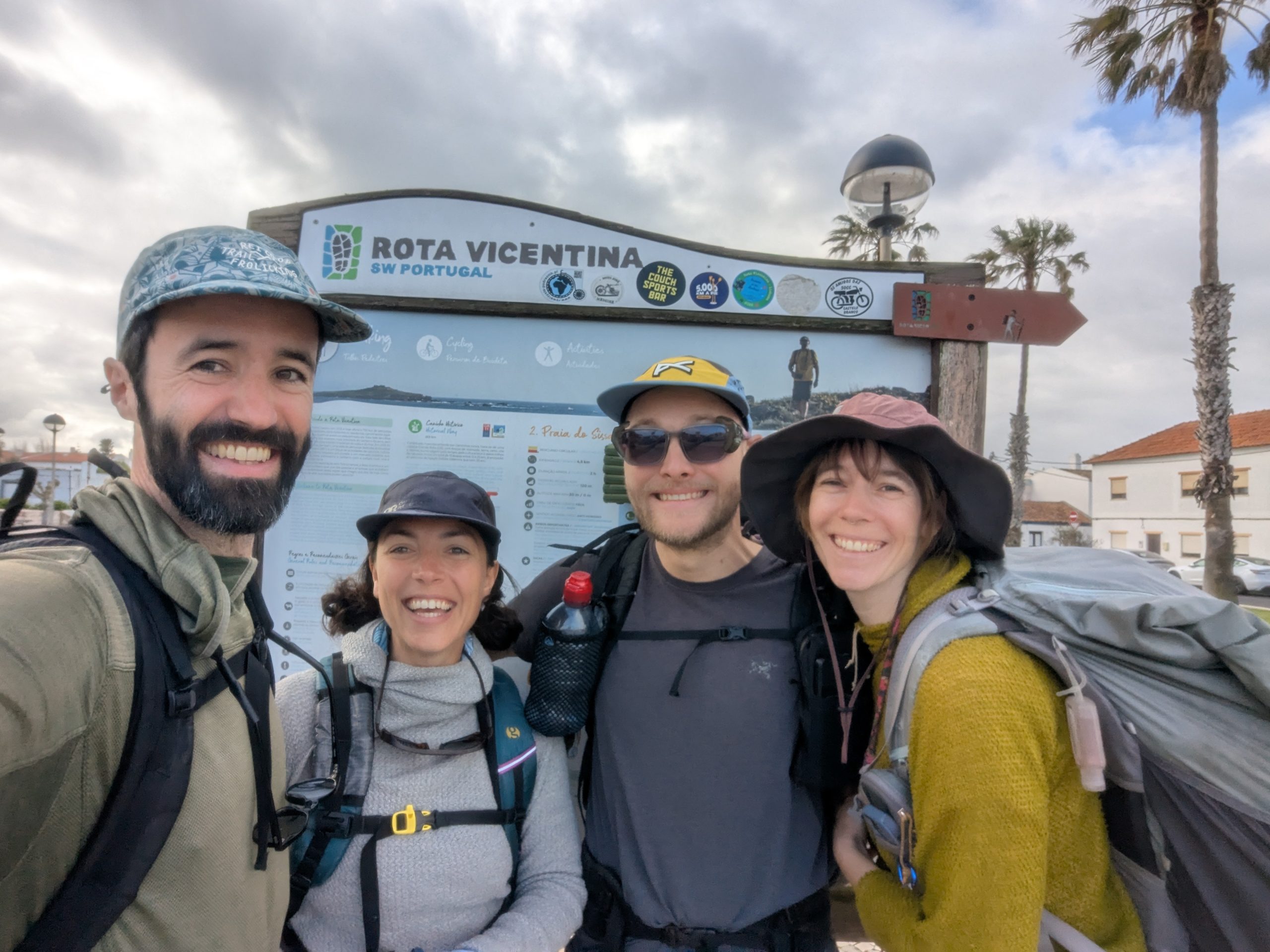 Four friends smiling in a selfie at the beginning of the Vicentina broken path, standing in front of the official sign of the trail on a sunny day.
