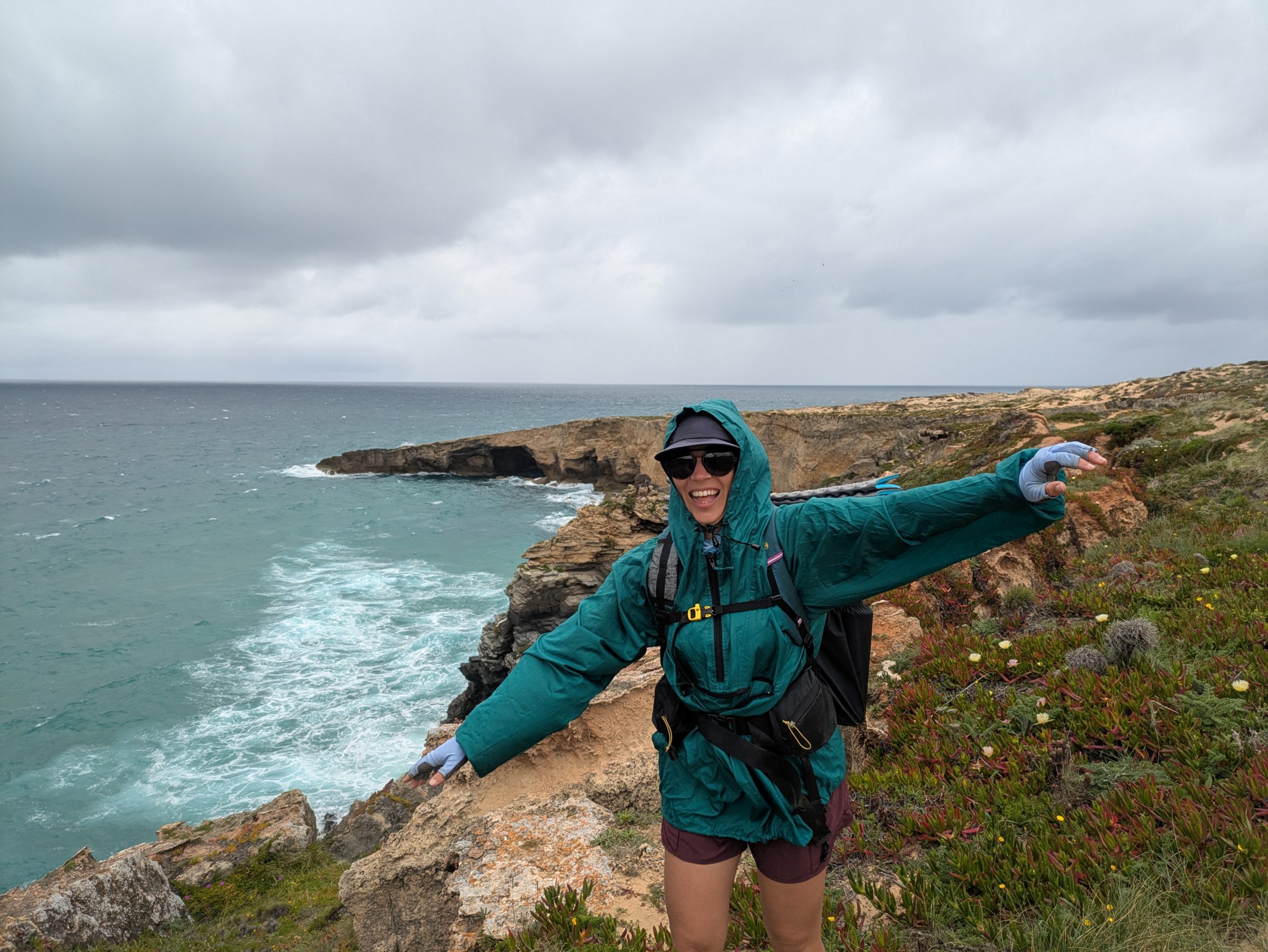 A hiker in a ground of wind, hood and flutter in the strong coastal wind, standing on a path with dramatic views of the ocean in the background.