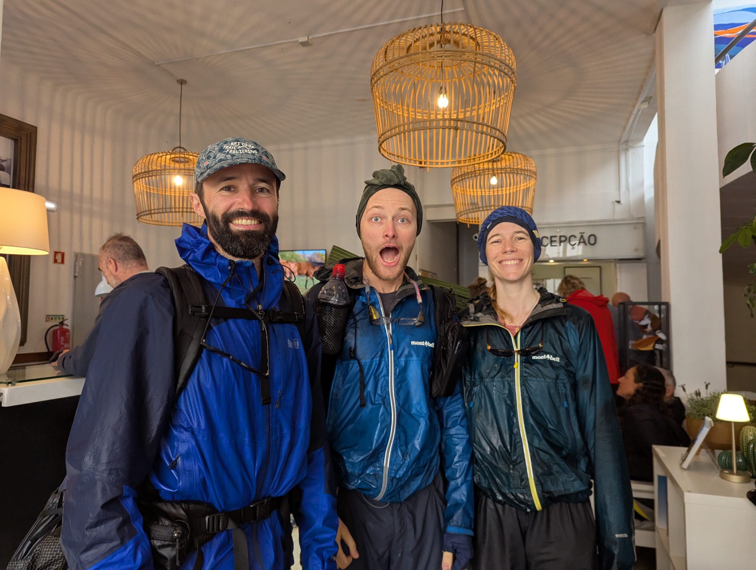 Three hikers standing in the area of reception of a hotel, soaked in the rain but smiling brilliantly. They carry wet rain equipment and look happy despite the weather.