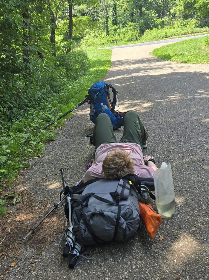Backpack woman lying on the pavement by grass with her head in her backpack.