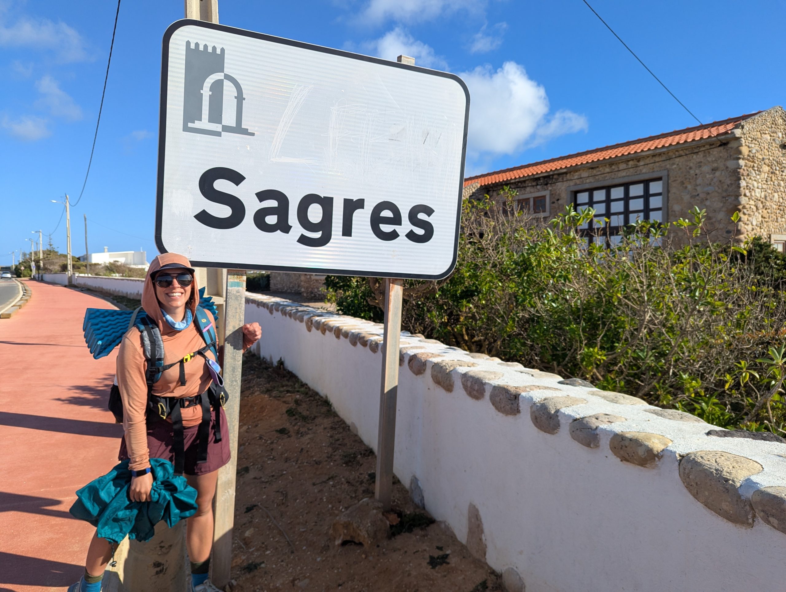 Chloe standing next to the Sagres sign after walking along the path of fishermen from Vila Do Bispo, smiling at the camera with a view to the ocean behind her.
