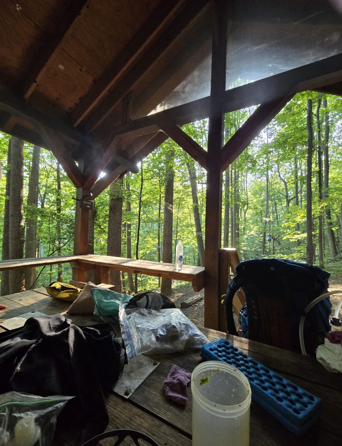 Excursion equipment scattered on a large picnic table under a porch roof.