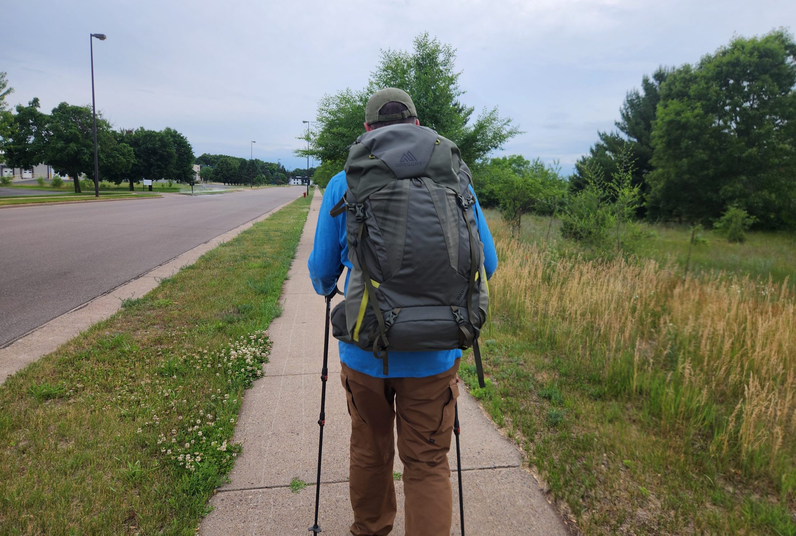 A hiker carrying a large Gregory Zulu backpack and trekking poles walks away from the camera along a suburban sidewalk, with an open road to the left, overgrown trees and grassland to the right.
