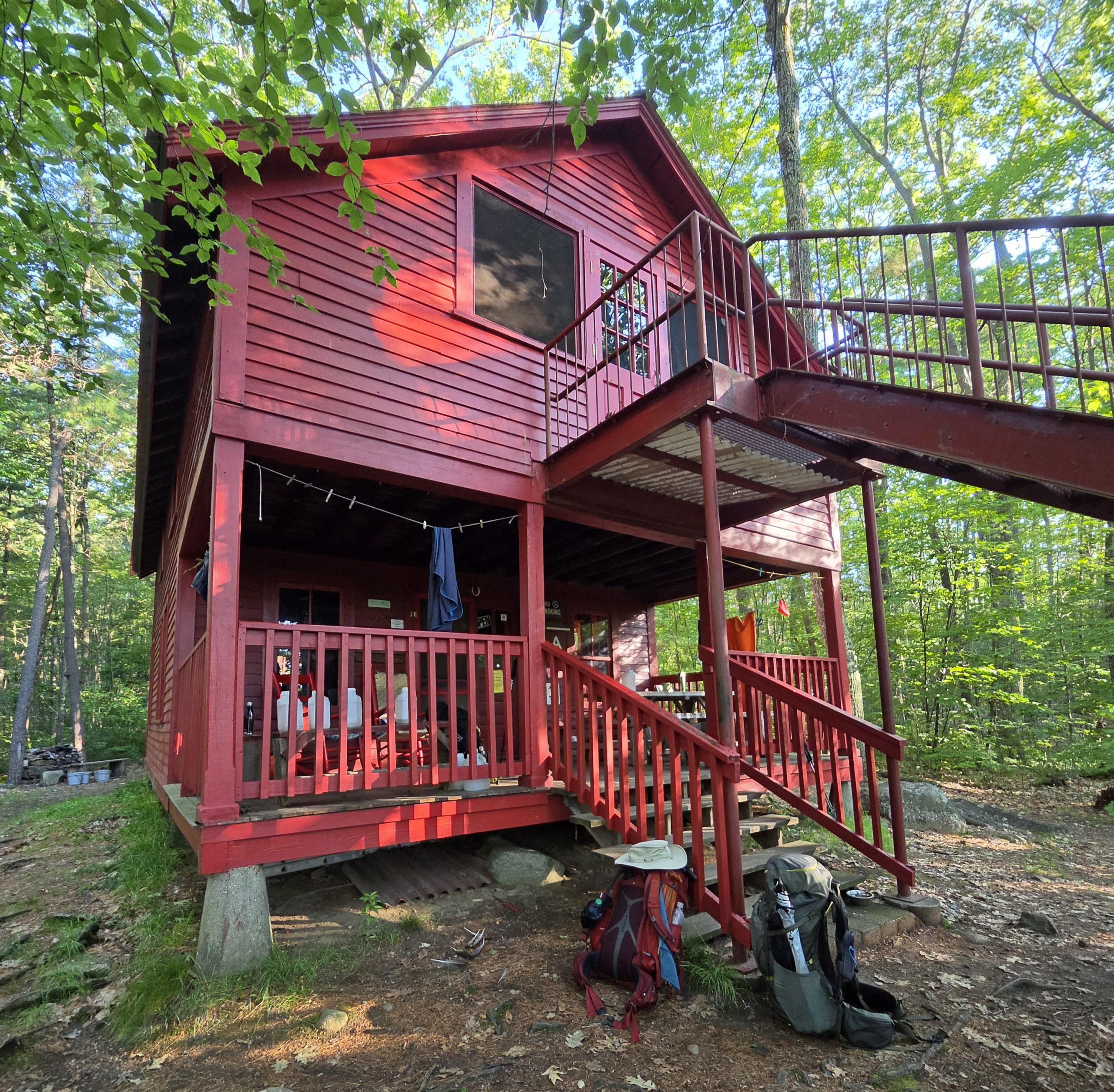 A two-story cabin with fresh barn red paint and a covered front porch, nestled among green trees, with rocking chairs and clotheslines on the porch.