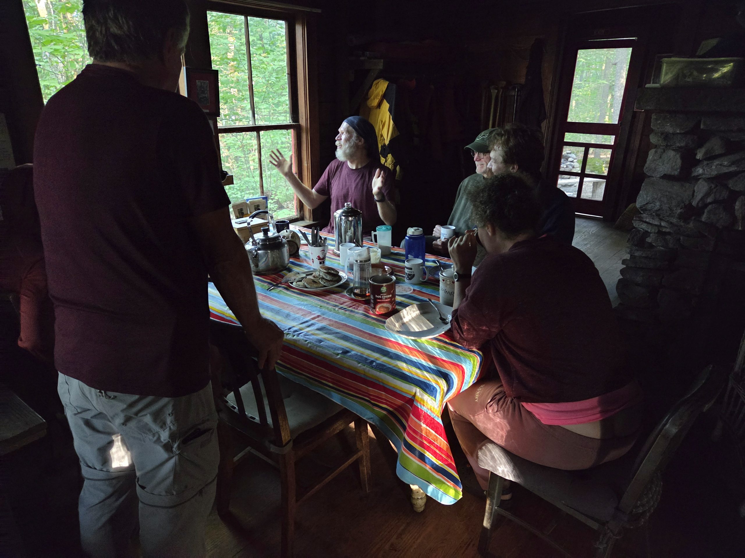Hikers sit around a table with a striped tablecloth inside a dim log cabin, sharing a breakfast of pancakes, coffee and hot chocolate, with green forests visible through the window.